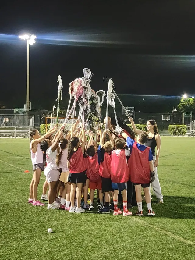 Redhawks players raise their lacrosse sticks together in a huddle at night under the field lights