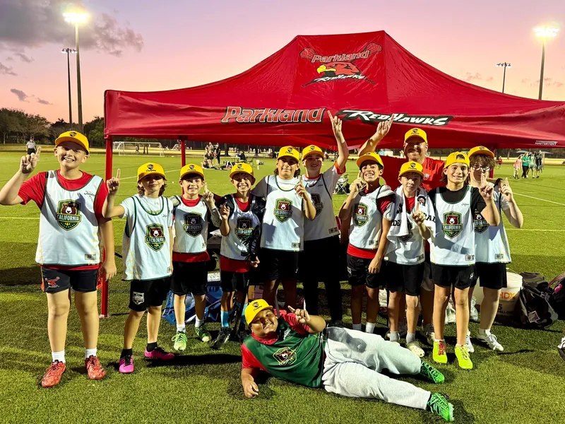 Young Redhawks players in white pinnies and yellow caps cheer together under the red Redhawks tent at dusk