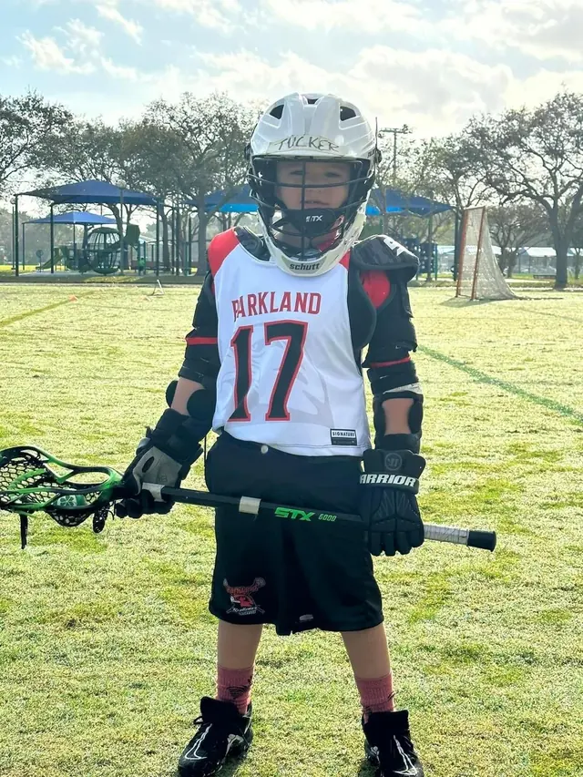 A young Redhawks boy in a helmet and Parkland jersey holds his lacrosse stick